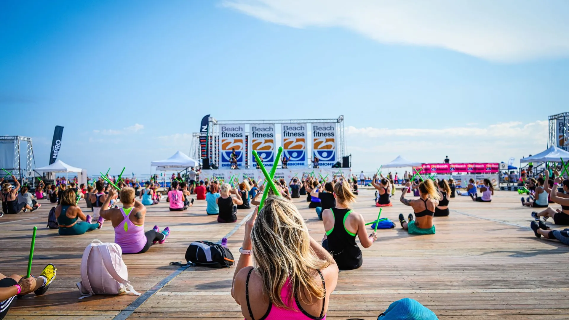 Güneşli bir havada ahşap bir platform üzerinde oturan, çoğu kadınlardan oluşan kalabalık bir grup, Beach fitness logolu sahneye dönük şekilde yeşil baget benzeri sopalarla ritmik bir açık hava etkinliğine katılarak enerjik bir spor turizm deneyimi sergiliyor.