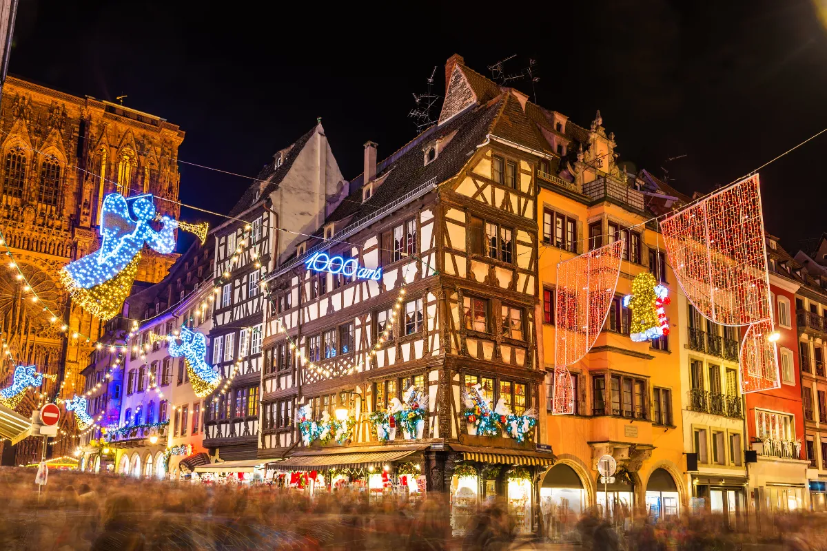 Strasbourg'un tarihi merkezinde, Noel'in Başkenti ruhunu yansıtan yarı ahşap evlerin ve Katedral'in süslü cephesinin önünde, ışıklı melek figürleri ve diğer bayram süslemeleriyle dolu, 2026 Avrupa Noel Pazarları atmosferinde kalabalık bir gece manzarası.