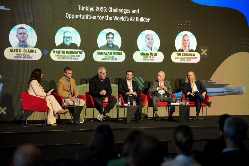 Bir konferans sahnesinde, Super Yacht Summit Türkiye etkinliğine ait büyük bir logo önünde, takım elbiseli beş erkek panelist masada oturmuş, önlerindeki mikrofonlarla canlı bir tartışma yürütüyorlar.