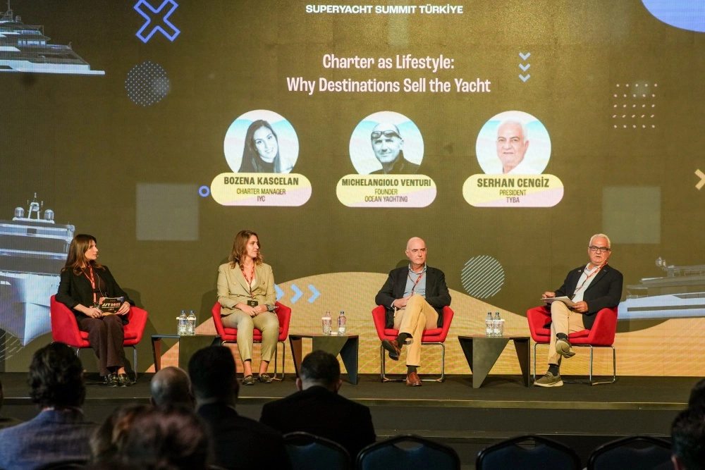 Türkiye Süper Yat Zirvesi konferans panelinde, parlak ışıklar altında bir masada oturan altı uzman, dikkatle dinleyen kalabalık izleyici önünde sektöre dair kritik konuları tartışıyor, her birinin önündeki mikrofonlar konuşmalarını kaydederken arkalarındaki büyük ekranda ilgili bir sunum görünüyor.