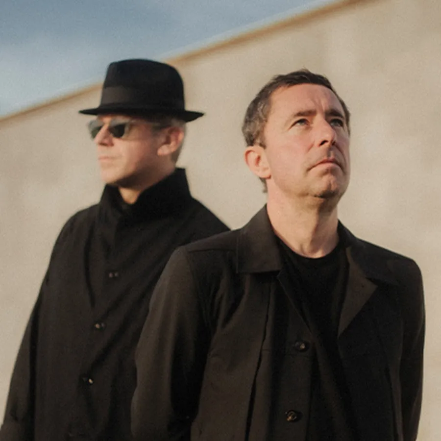 The Cinematic Orchestra grubundan iki üye, açık renkli bir arka plan önünde, siyah paltolarıyla görülüyor; sağdaki üye başını yukarı kaldırıp düşünceli bir ifadeyle bakarken, soldaki üye şapka ve güneş gözlüğüyle arkasında duruyor.