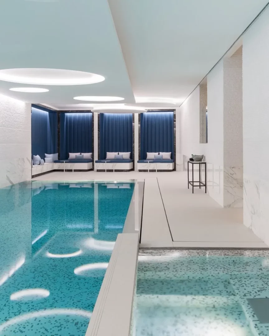 The Woodward Auberge'in lüks kapalı havuzunda, tavan ışıklarının suya yansıdığı açık mavi mozaik karolarla kaplı berrak su ve yan tarafta kadife mavi döşemeli üç adet dinlenme köşesi modern ve davetkar bir atmosfer sunuyor.