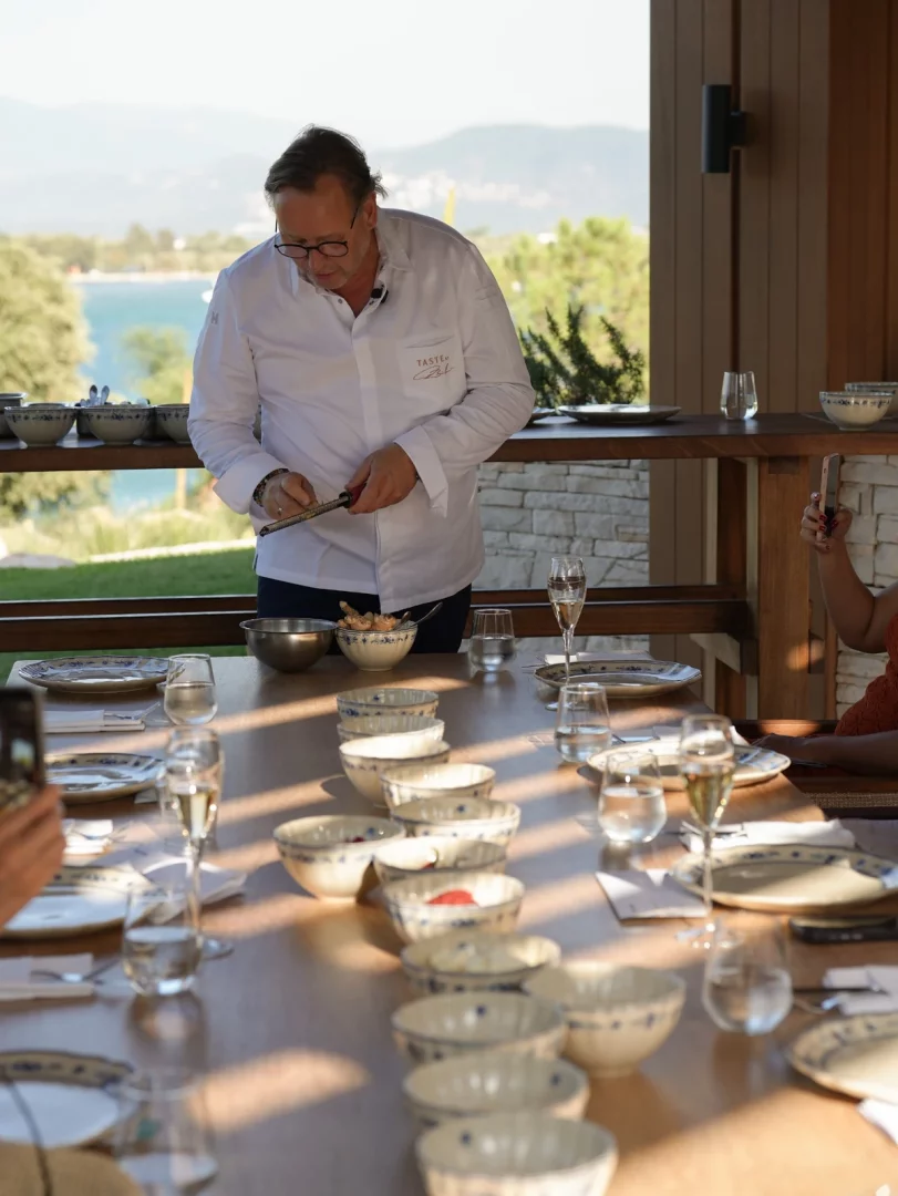 Şef Thomas Bühner, deniz ve dağ manzaralı açık hava bir ortamda düzenlenen yemek atölyesinde, beyaz şef ceketini giymiş bir şekilde uzun ahşap masada özenle malzeme rendeleyerek katılımcılara sunum yaparken, masada çok sayıda tabak, kase ve içecek kadehleri sıralanmıştır.