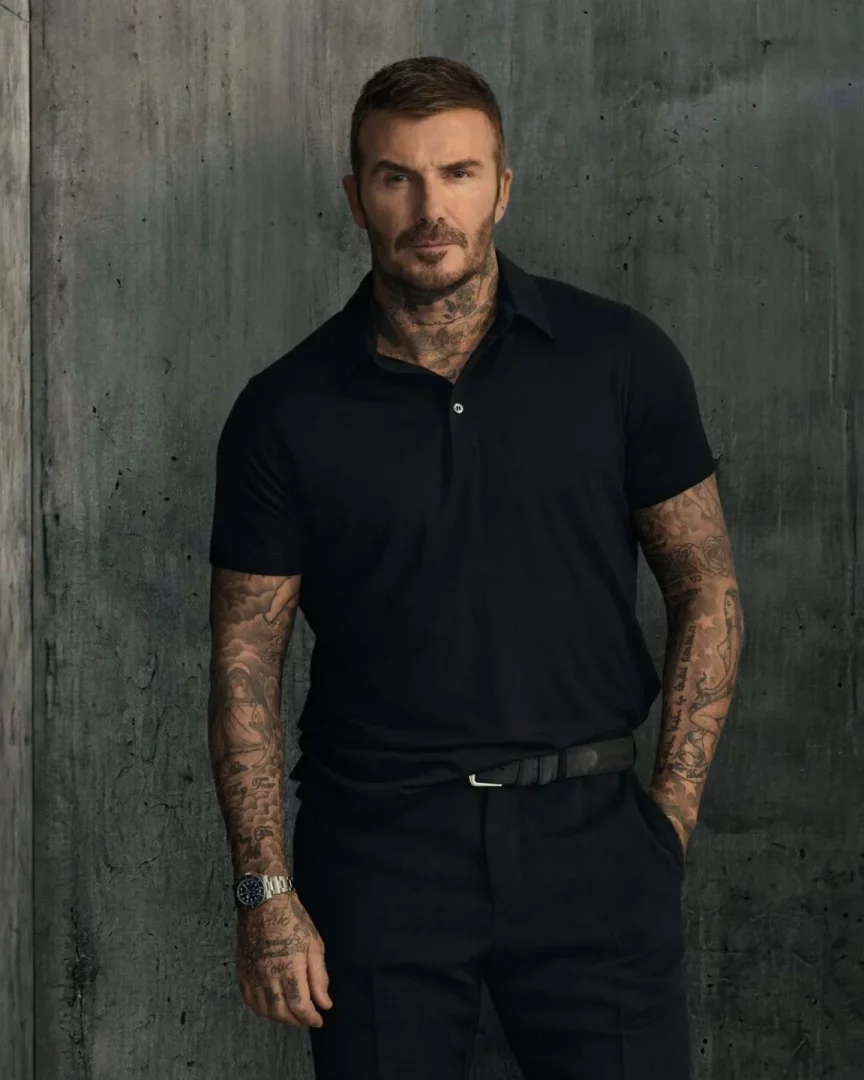 Koyu gri, dokulu bir arka planın önünde duran, siyah polo yaka tişört ve pantolon giyen David Beckham, boynu ve kolları tamamen dövmelerle kaplı bir şekilde, sol bileğindeki metal kayışlı lüks saatiyle kameraya ciddi bir ifadeyle bakıyor.