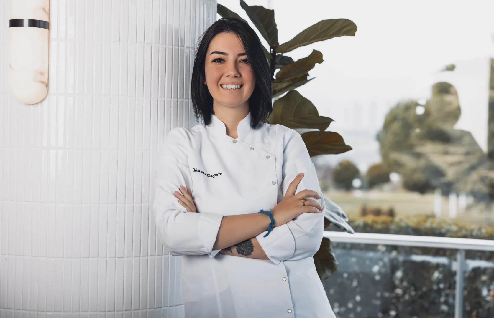 Şef Yaren Çarpar, kolları bağlı şekilde beyaz aşçı ceketiyle gülümseyerek bembeyaz fayans duvarın önünde duruyor; incir mevsimi boyunca menüsünde incire özel yer veren şeflerden biri olarak öne çıkıyor.