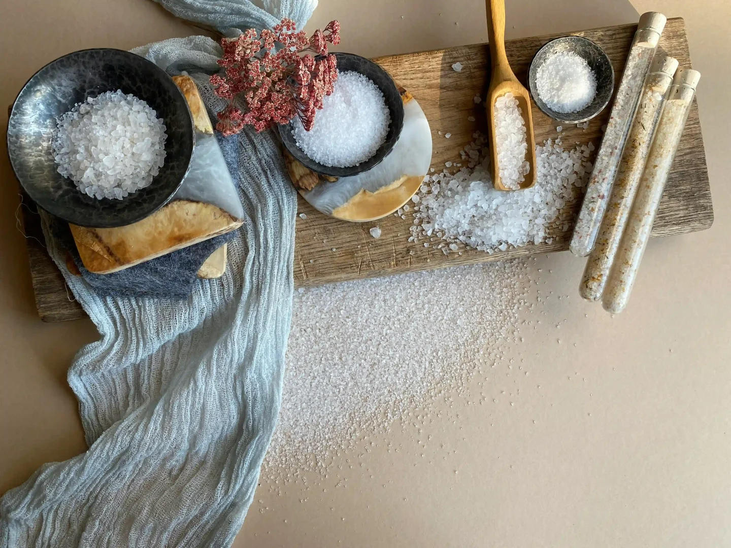 Ahşap bir kesme tahtası üzerinde, iki koyu renk kasede iri taneli, küçük bir gümüş kapta ve ahşap kaşıkta farklı tuz çeşitleri ile mantar tıpalı üç cam tüpteki aromalı tuzlar, açık mavi dokulu kumaş ve bej zemine dağılmış tuz kristalleriyle birlikte estetik bir düzenleme oluşturuyor.