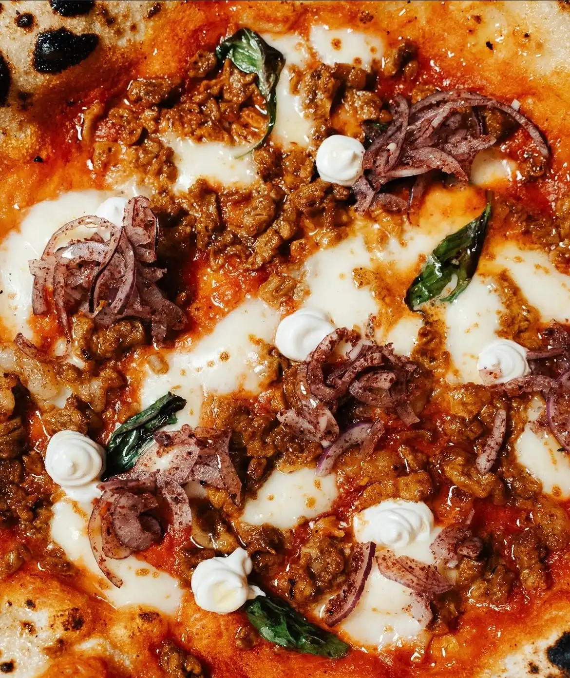 Bu yakın çekim pizza görüntüsünde, Çengelköy'deki bir mekandan alınan, nar gibi kızarmış kenarlı hamuru üzerinde erimiş beyaz peynir, kavrulmuş kıyma, ince dilimlenmiş kırmızı soğan, taze fesleğen ve küçük kremalı sos dokunuşlarıyla zenginleştirilmiş iştah açıcı bir pizza detayı öne çıkıyor.