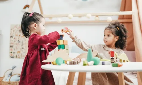 Kırmızı elbiseli, koyu düz saçlı bir kız ile açık renk giysili, kıvırcık saçlı diğer bir kız çocuğunun, bir masanın etrafında ahşap bloklar ve eğitici oyuncaklarla neşeyle oynayarak bilişsel becerilerini ve beyin gelişimlerini güçlendirdiği görülüyor.