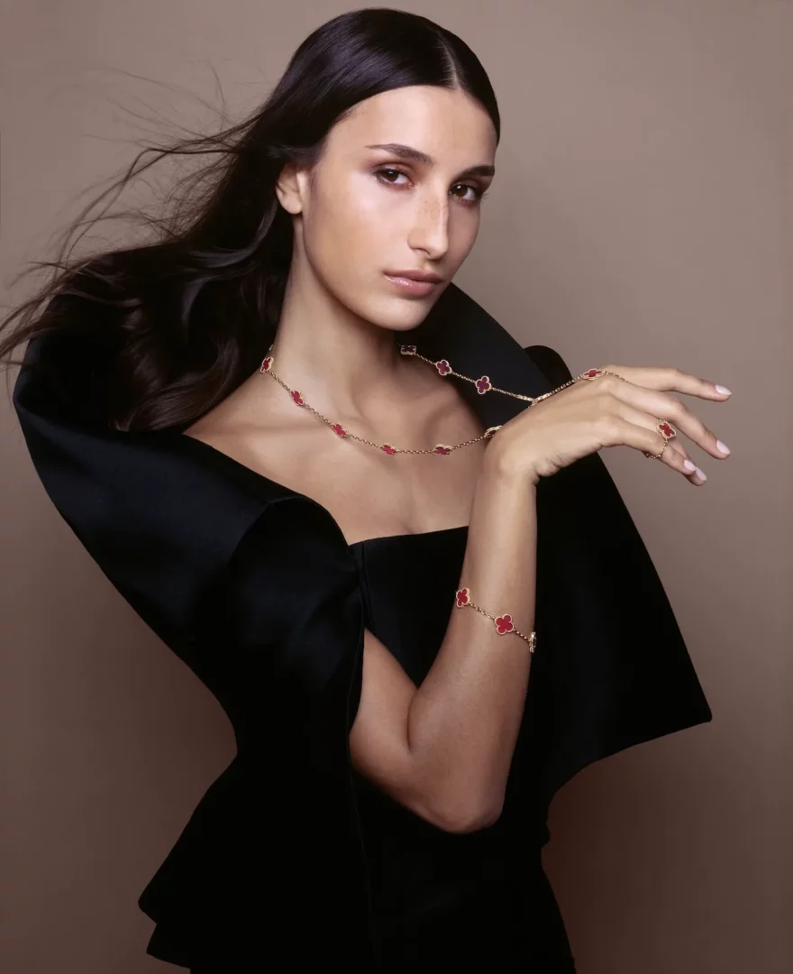 Siyah zarif bir kıyafet giyen ve rüzgarda hafifçe savrulan saçlara sahip genç bir kadın, boynunda, bileğinde ve parmağında Van Cleef & Arpels'in ikonik kırmızı yonca motifli Alhambra mücevher setini sergiliyor, zarafeti ve şıklığı vurguluyor.