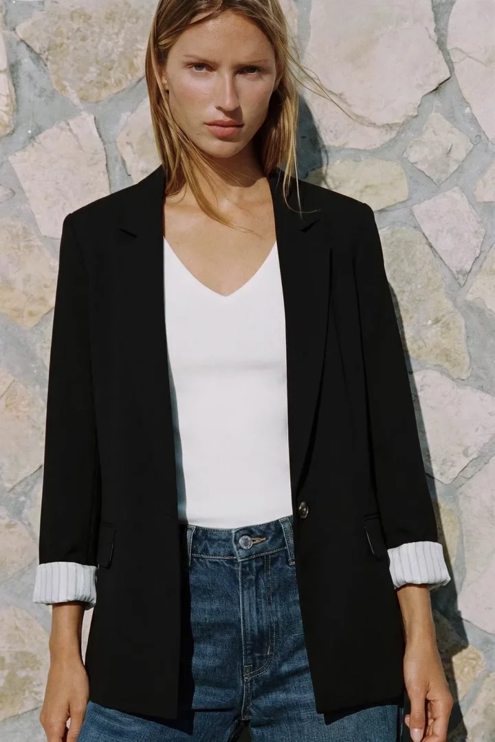 Taş duvar fonunda, üzerinde şık bir siyah blazer ceket, beyaz V yaka bir üst ve mavi kot pantolon bulunan, manşetleri katlanmış ceketin çizgili astar detayını belirgin şekilde gösteren, uzun saçlı genç bir kadın modern ve tarz bir görünüm sergiliyor.