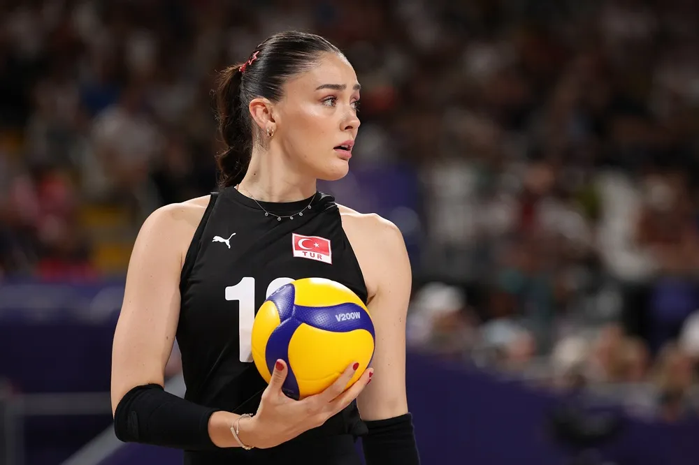 Zehra Güneş, siyah milli takım formasının üzerinde Türkiye bayrağı ve 18 numarasıyla, sarı-mavi voleybol topunu sol elinde tutarken maça odaklanmış bir şekilde sağa bakıyor; arka planda seyircilerle dolu bir spor salonu bulanık bir şekilde görülüyor ve bu an, Filenin Sultanları ruhunu yansıtıyor.