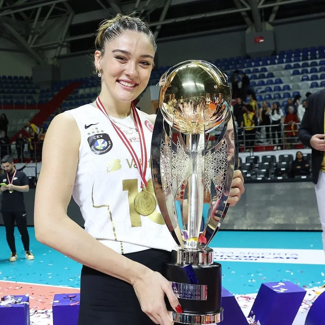 VakıfBank formasıyla, boynunda kırmızı kurdeleli madalyalar ve elinde parıldayan büyük şampiyonlar kupasıyla zaferle gülümseyen voleybolcu Zehra Güneş, tribünlerin ve sahnedeki konfetilerin arasında, kazandığı ödüllerle gururla poz veriyor.