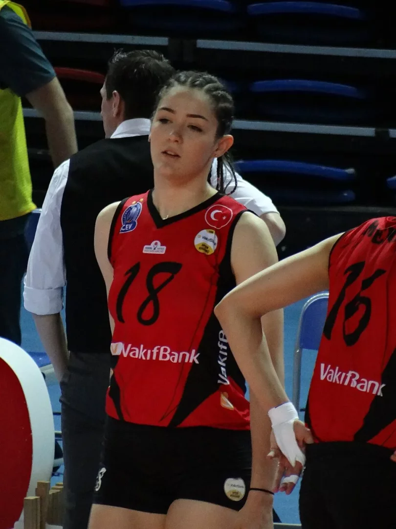 Zehra Güneş, VakıfBank formasıyla 18 numaralı kırmızı ve siyah voleybol tişörtü giymiş, saçları örgülü bir şekilde sahada duruyor; arkasında bulanık tribünler ve diğer takım üyeleri görünürken, bu an voleybol kariyerine nasıl başladığına dair hikayesiyle bütünleşiyor.