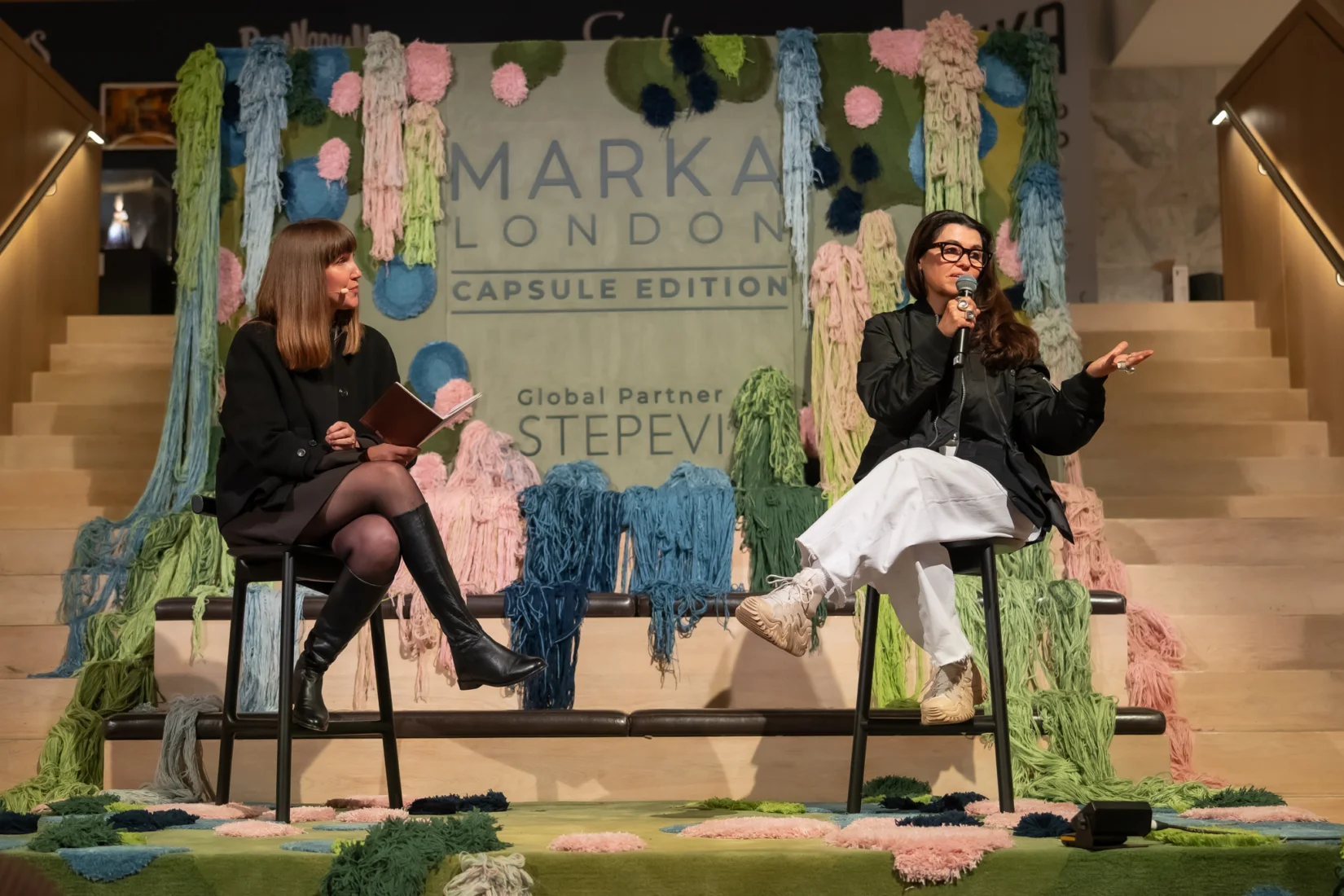 MARKA London konferansında, sahnedeki konuşmacılardan sanatçı ve tasarımcı Es Devlin'in mikrofon başında izleyicilere seslendiği, yanında moderatör Johanna Agerman Ross'un ise dikkatle dinlediği, katılımcılara yeni ve ilham veren fikirler sunan bir an.