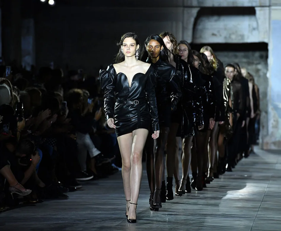 Bir Saint Laurent defilesinde, Anthony Vaccarello'nun modaevindeki 10 yıllık dönemine damgasını vuran keskin ve cüretkar tarzını yansıtan, derin V yaka ve pofuduk kollu siyah deri mini elbise giyen bir model, arkasında diğer modellerle podyumda yürüyor, izleyiciler karanlık ortamda onları izliyor.