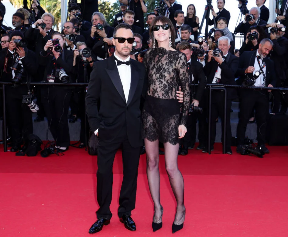 Anthony Vaccarello, siyah smokini ve güneş gözlükleriyle, Saint Laurent’ın unutulmaz kreatif direktörlüğü döneminin ruhunu yansıtan transparan siyah dantelli elbisesiyle Charlotte Gainsbourg ile kırmızı halıda poz veriyor, arka planda fotoğrafçılar flaşlarını patlatıyor.