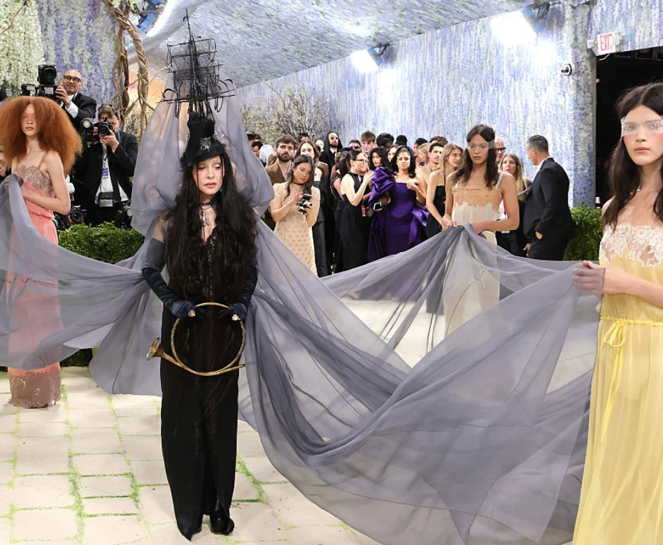 Anthony Vaccarello tasarımı Saint Laurent imzalı siyah dantel elbisesiyle 2026 Met Gala'da boy gösteren Madonna, gemi biçimli şapkası, uzun eldivenleri ve elindeki altın boynuzla dikkat çekerken, kıyafetini tamamlayan uçuşan gri pelerini etrafındaki modeller tutuyor.