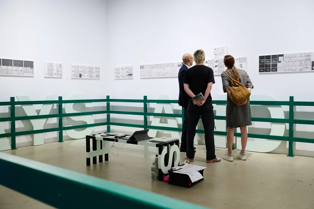 Büyük bir sanat galerisinde, arkası dönük bir izleyici, Art Basel gibi önemli bir sanat etkinliğinin atmosferini yansıtan, kırmızı, turuncu, sarı ve mavi tonlarındaki canlı renklerle dolu, güçlü fırça darbelerine sahip soyut bir tabloya odaklanmış bir şekilde bakıyor.