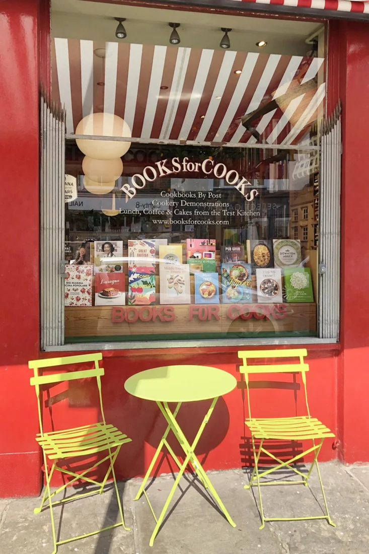 Kırmızı cepheli Books for Cooks adlı gastronomi kitapçısının vitrininde, beyaz-kırmızı çizgili tente altında dünya mutfaklarından tarifler içeren çeşitli yemek kitapları sergileniyor; önündeki parlak lime yeşili masa ve iki sandalye, raflardaki lezzetlerden ilham alarak keyifli bir mola için ziyaretçilerini bekliyor.