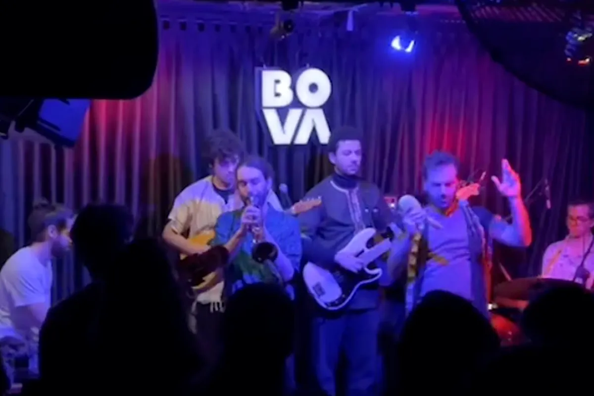 Beyoğlu'ndaki Bova Sahne'nin canlı caz performansında, basçı, trompetçi ve vokalistten oluşan bir müzik grubu, mekanın aydınlatmalı BOVA logosu önünde mavi ve kırmızı sahne ışıkları altında çalarken, dinleyiciler İstanbul'un en iyi caz kulüplerinden birindeki müziğin keyfini çıkarıyor.