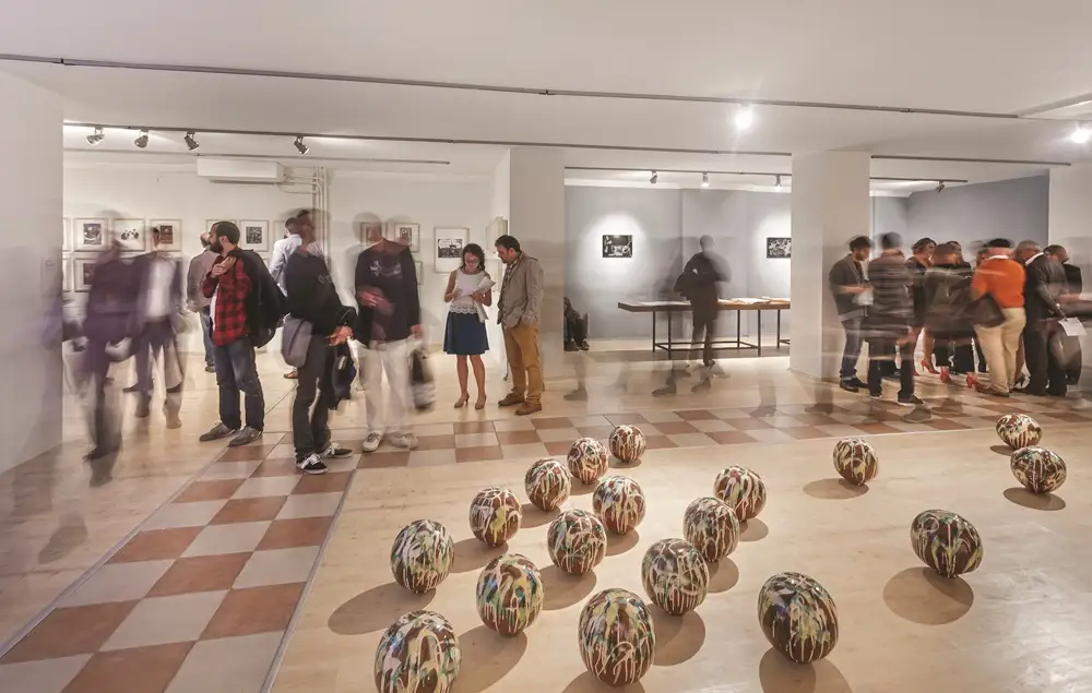 Çanakkale Bienali sergisinden bir karede, Ege Denizi'nin kıyısında, gün batımının sıcak ışıkları altında duran, çağdaş sanatın özgün bir ifadesi olan büyük ölçekli ve geometrik formlara sahip bir heykel enstalasyonu, çevresindeki ziyaretçilerin dikkatini çekiyor.