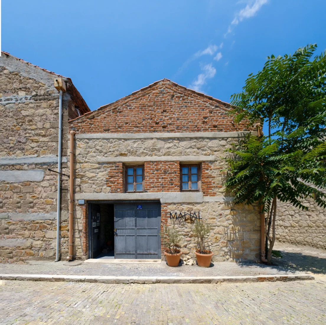 Çanakkale Bienali'ndeki Mahal adlı sergi alanında, metal iskeletli bir enstalasyonun önünde gri tonlarda büyük soyut bir duvar resmi ve organik formlarda heykellerin sergilendiği modern bir iç mekan görünümü.