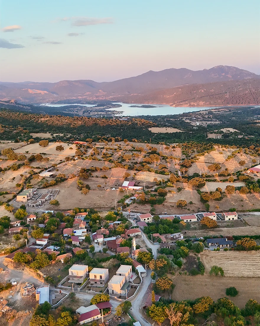 Çanakkale Bienali kapsamında Tartışık Koyu'nun etkileyici doğal peyzajını gözler önüne seren bu geniş açılı görselde, güneşin aydınlattığı açık mavi denizin sakin suları kayalık ve çakıllı bir kıyıya nazikçe vururken, ufukta yükselen seyrek bitki örtüsüyle kaplı yeşil tepeler gökyüzüyle huzurlu birleşiyor.