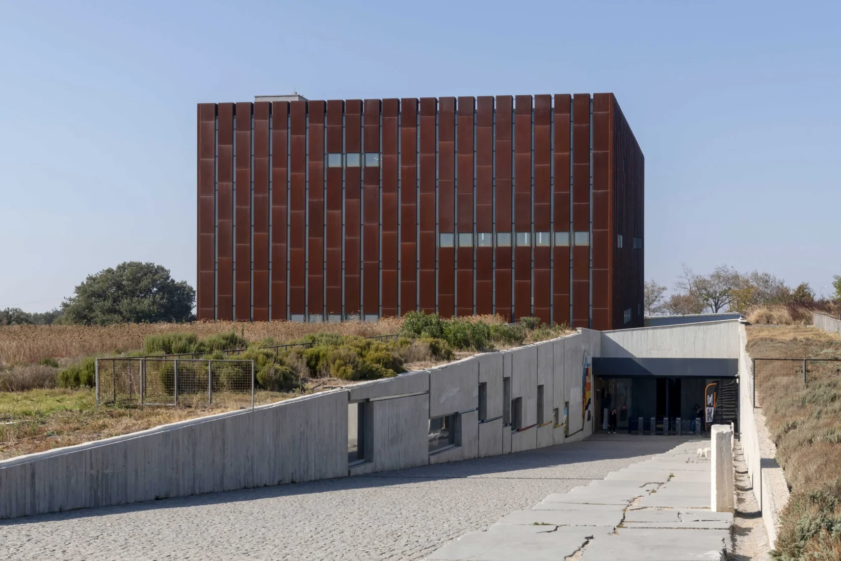 Üçgenimsi keskin hatlara sahip, modern ve gri tonlardaki Troya Müzesi'nin etkileyici dış cephesi, bulutlu gökyüzünün altında Çanakkale Bienali'nin ruhunu yansıtarak ön planda gururla dalgalanan bir Türk bayrağı ile dikkat çekiyor, kültürel miras ile güncel sanatı birleştiriyor.