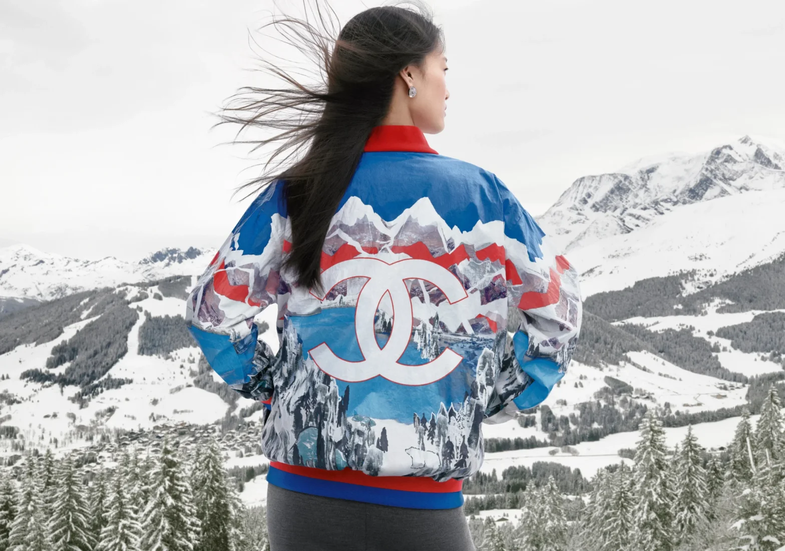 Karlı Fransız Alpleri'nin geniş manzarasına sırtı dönük bakan bir model, Chanel Coco Neige 2026 koleksiyonunun canlı renklerdeki, arkasında dağ, göl ve kutup ayısı detaylı bir Kayak Modası bomber ceketini giyiyor, ceket üzerindeki büyük beyaz Chanel logosu dikkat çekiyor.