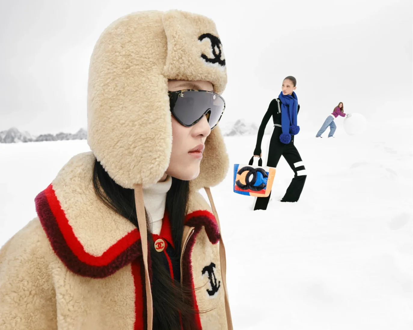 Ön planda kabarık bej bir kulaklıklı şapka ve büyük güneş gözlükleri takan model, karlı Megève manzarasında, geride renkli Chanel logolu bir çanta taşıyan başka bir model ve kar topu yuvarlayan bir kadınla, Chanel Coco Neige 2026'nın retro kayak modasına getirdiği sportif ve neşeli havayı gözler önüne seriyor.