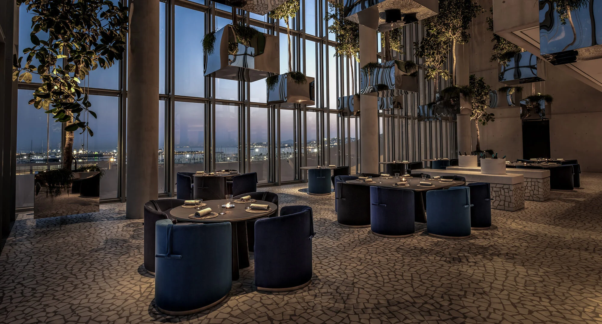 Delta Restaurant'ın şık iç mekanında, beyaz örtülü masalar özenle hazırlanmış servis takımları ve kadehleriyle misafirlerini beklerken, loş ve sıcak ışıklandırma modern ahşap detaylarla birleşerek dışarıdaki şehir ışıklarını yansıtan büyük camların ardında davetkar bir atmosfer sunuyor.
