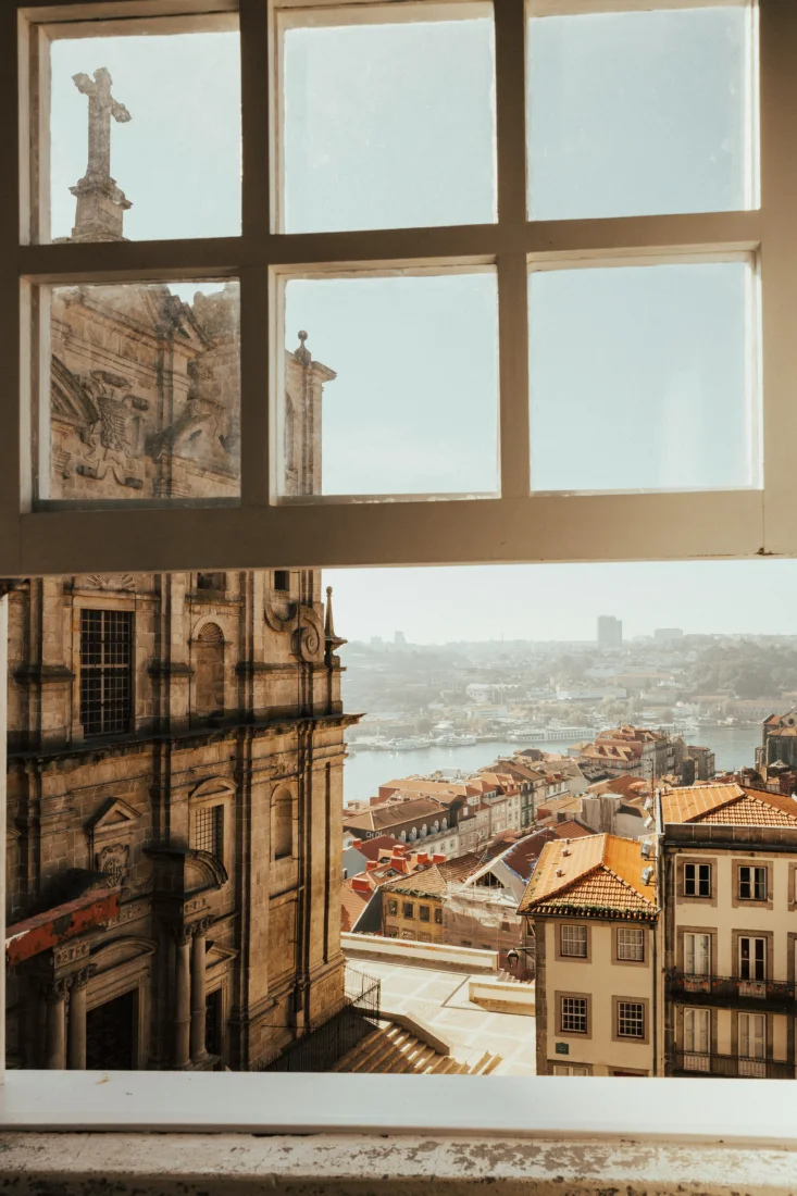 Pencere çerçevesinden görünen, güneşli bir havada Douro Nehri kıyısındaki tarihi Porto şehrinin, yüksekte duran haç figürü, ayrıntılı taş işçiliğine sahip eski yapıları ve portakal renkli çatılı evleriyle Sonbahar-Kış ayları için belirlenen mikro mevsim rotalarında keşfedilecek kültürel zenginliğini yansıtan panoramik manzarası.