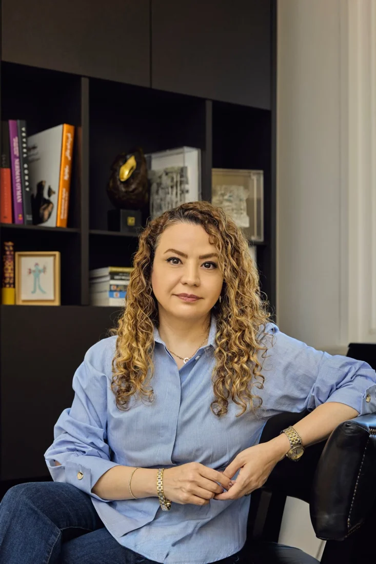 Zarifçe gülümseyen, koyu saçlı Elnaz Rahimoğlu'nun yakın çekim portresi; omuzdan yukarısı görünen, koyu ve dokulu bir kıyafetle, gözleri hafifçe kadraj dışına dönük, yumuşak ışıklandırmanın vurguladığı detaylarla kendinden emin ve şık bir duruş sergiliyor.
