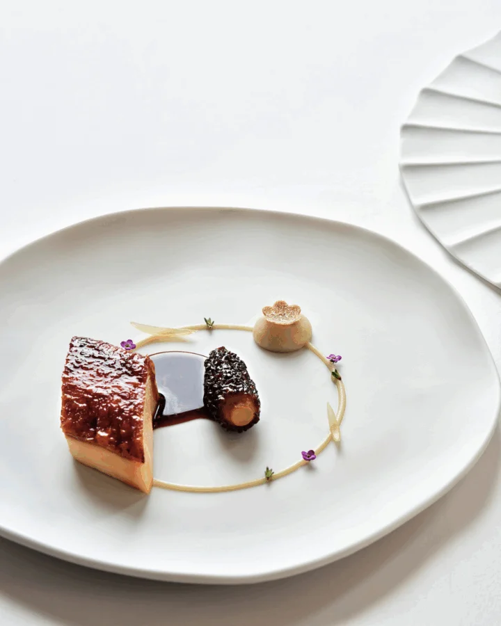 Beyaz dikdörtgen bir tabakta, özenle yerleştirilmiş küçük porsiyon bir ana yemek, ince soslar ve taze yeşilliklerle sanatsal bir sunumla servis ediliyor, bu da üst düzey bir fine dining deneyiminin gastronomik estetiğini ve lezzet arayışını vurguluyor.