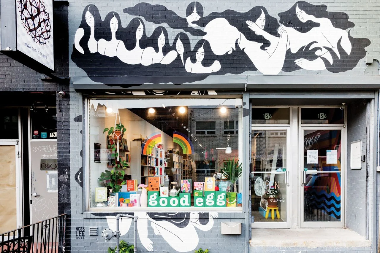 Gri tuğla duvarlı bir binanın dış cephesinde, uzun saçlı soyut kadın figürlerinin yer aldığı siyah beyaz bir duvar resmi bulunan Toronto'daki 'good egg' adlı gastronomi kitapçısının geniş cam vitrininde, raflar dolusu yemek kitabı, renkli mutfak aksesuarları ve tasarım objeleri sergileniyor, böylece burası tariflerden çok daha fazlasını sunuyor.