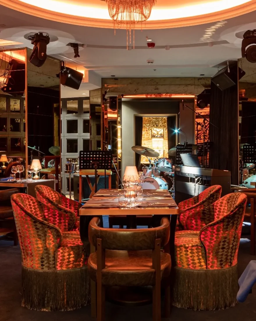İstanbul'un en iyi caz kulüplerinden Gregor Jazz Club'ın sıcak ve samimi atmosferini gözler önüne seren bu fotoğrafta, sahnedeki davul seti ve klavye, yansımalı duvarlar ve zarif avize ile aydınlatılan, konforlu masa düzenlemeleri ve kırmızı desenli koltuklar, canlı caz performansları için ideal bir ortam sunuyor.