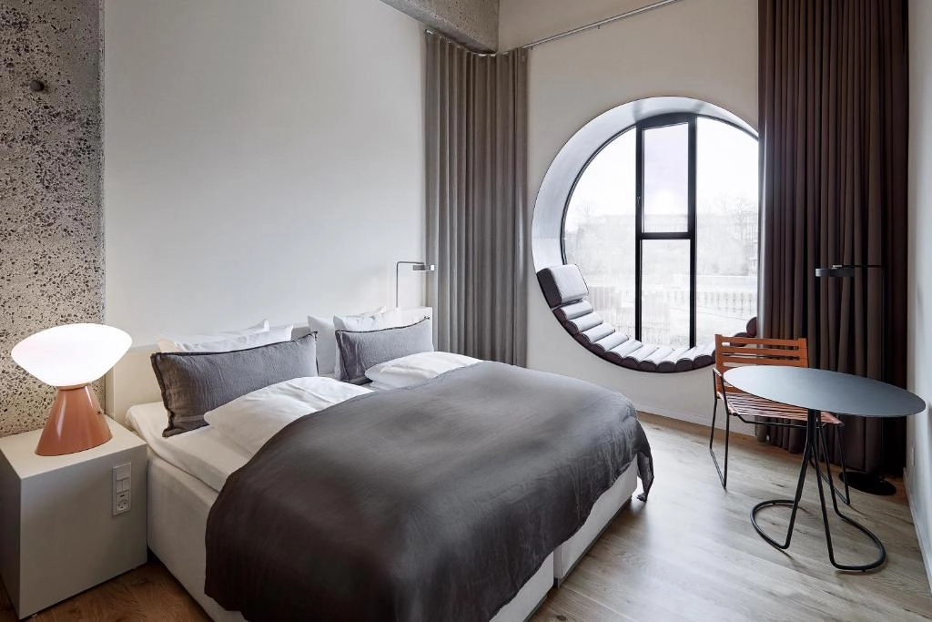 Hotel Ottilia'nın modern ve ferah odasından bir görünüm; büyük yatağı, minimalist komodinleri, kendine özgü aydınlatmaları ve şehir manzaralı geniş penceresiyle şık ve konforlu bir konaklama deneyimi vadediyor.