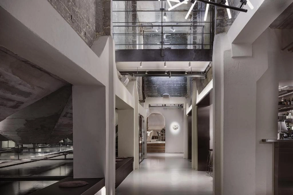 Hotel Ottilia'nın dikkat çekici tasarımına sahip modern iç mekanında, loş ışıklar altında uzun ahşap bir bar masasının etrafına yerleştirilmiş, zarif hatlara sahip tabureler, hem rahatlama hem de keyifli sohbetler için ideal, sofistike bir atmosfer yaratıyor.