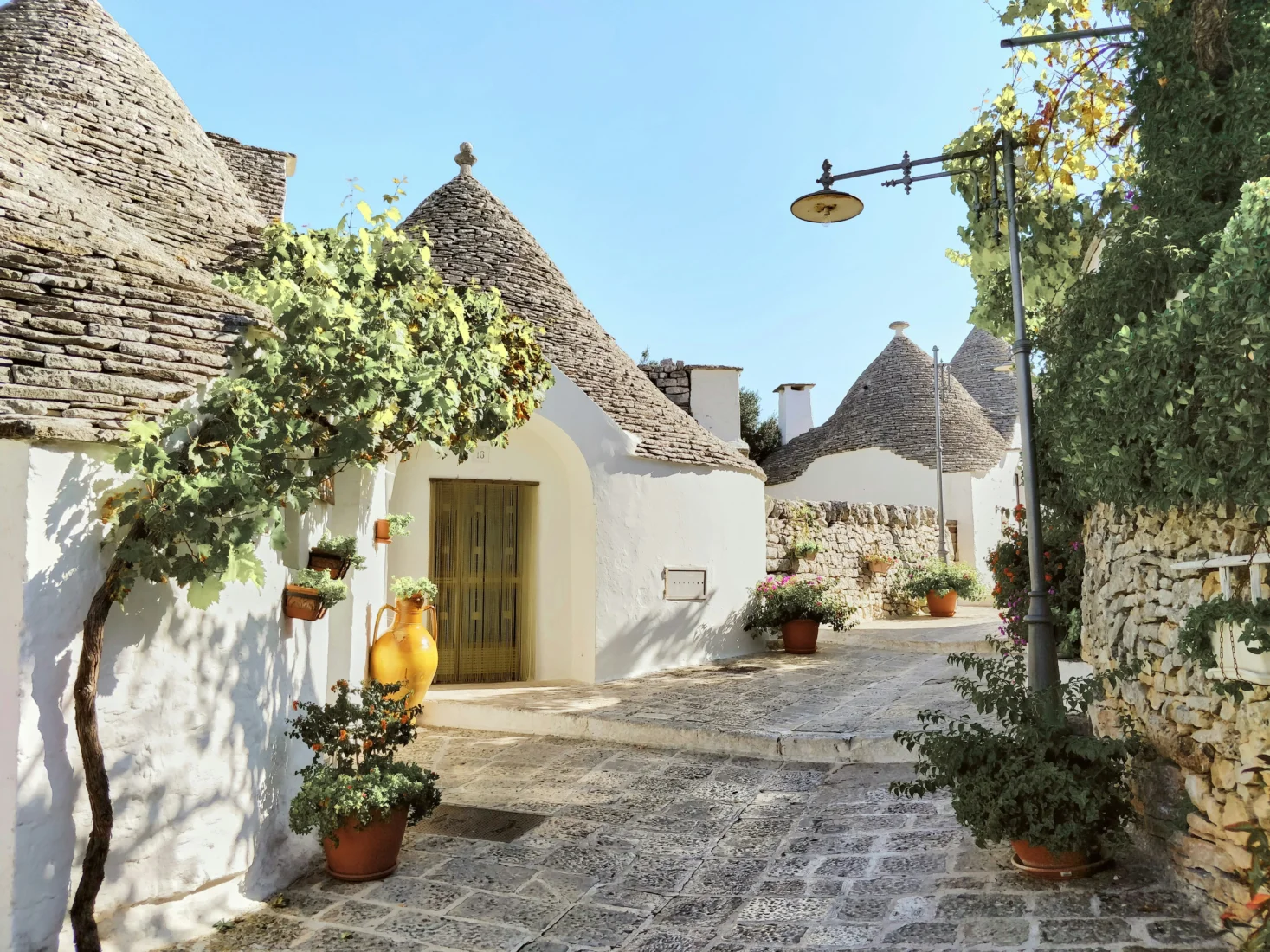 İtalya'nın Puglia bölgesindeki beyaz duvarlı ve konik taş çatılı trullo evlerinin sıralandığı, saksı çiçekleri ve yeşil sarmaşıklarla süslü Arnavut kaldırımlı sakin bir sokak, sonbahar-kış ayları mikro mevsim rotaları için eşsiz bir dinginlik sunuyor.