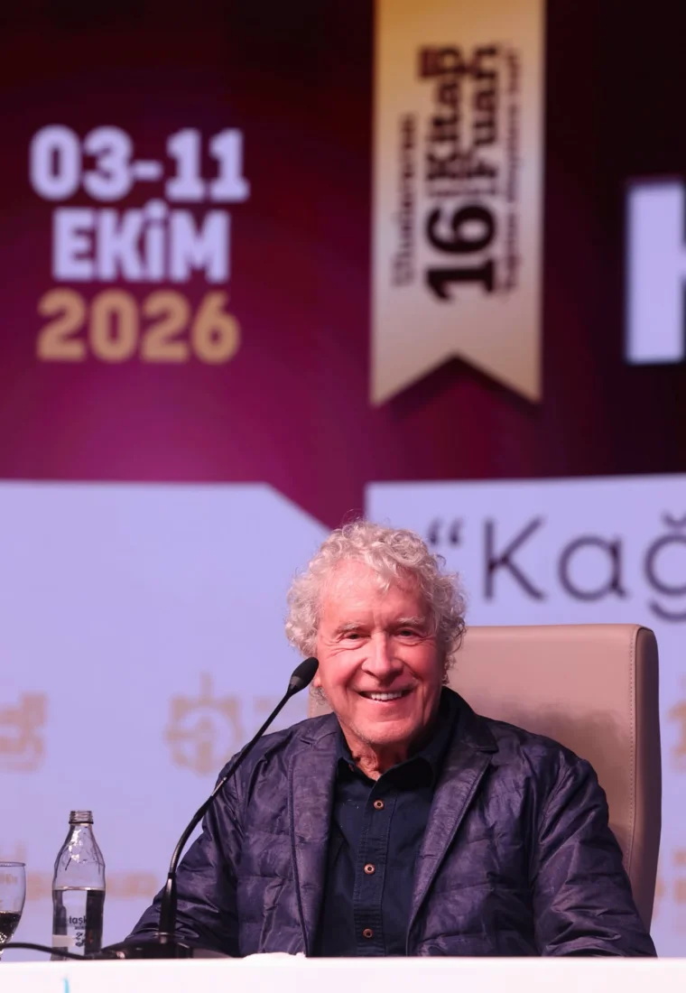 John Perkins, bir etkinlik sahnesinde, mikrofonun başında, derinlemesine düşünülmüş bir konu hakkında konuşurken, odaklanmış ve deneyimli bir konuşmacı edasıyla dinleyicilere bakıyor, küresel ekonomik ve siyasi sistemler üzerine yaptığı analizlerle bilinen kişiliğini yansıtıyor.