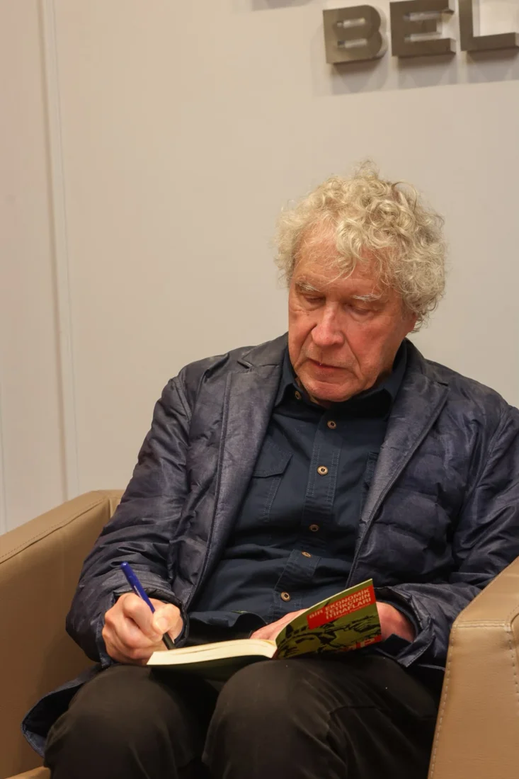 Beyaz saçları ve sakalıyla dikkat çeken, tanınmış yazar John Perkins, gri çerçeveli gözlüklerinin ardından doğrudan kameraya odaklanmış, derinlemesine düşüncelerini ve yaşam deneyimlerini yansıtan bilge ve sakin bir ifadeyle poz veriyor.