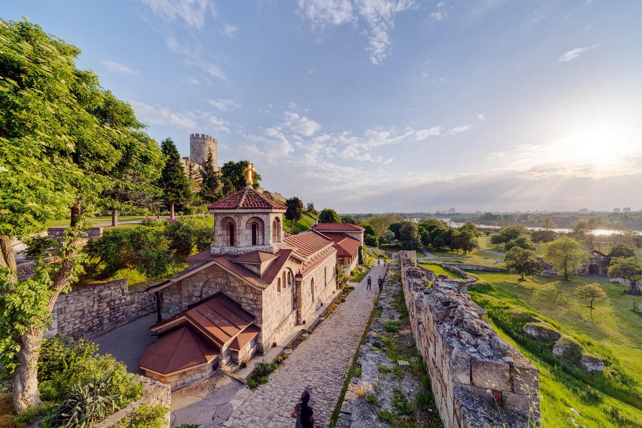 Belgrad'ın kalbinde yer alan, yemyeşil ağaçlarla çevrili tarihi Kalemegdan Parkı'nın taş surları arasında yürüyen ziyaretçiler ve uzaktaki şehir manzarasıyla dolu, dingin bir anı yakalayan geniş bir fotoğraf.