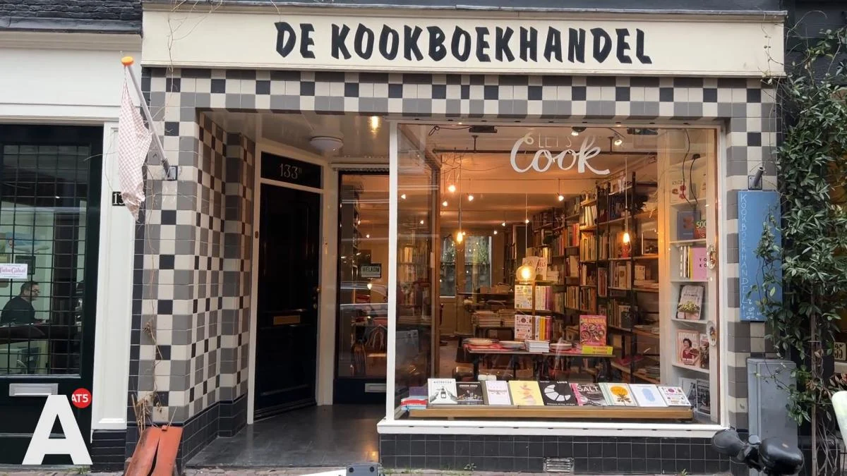 Amsterdam'da yer alan, yüksek tavanları ve katlara uzanan ahşap rafları kitaplarla dolu, birkaç kişinin sakin bir şekilde dolaştığı, etkileyici ve geniş bir kitapçı mekanının iç görünümü.