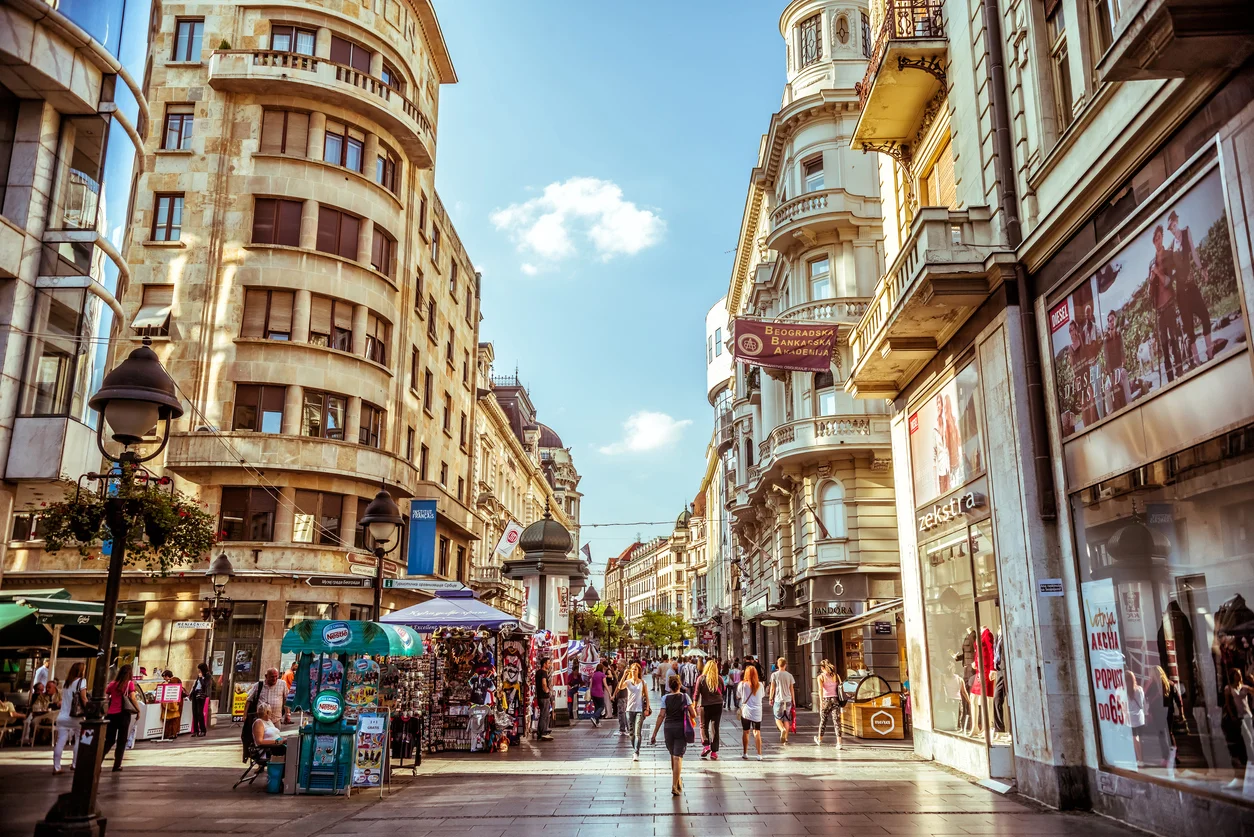 Knez Mihailova Caddesi'nin güneşli bir öğleden sonrasında, cadde boyunca sıralanmış tarihi mimariye sahip çok katlı binaların zemin katlarındaki mağaza ve kafelerin önünde, ağaçların gölgesinde yürüyen çok sayıda insanın oluşturduğu hareketli ve canlı bir şehir hayatı görüntüsü.
