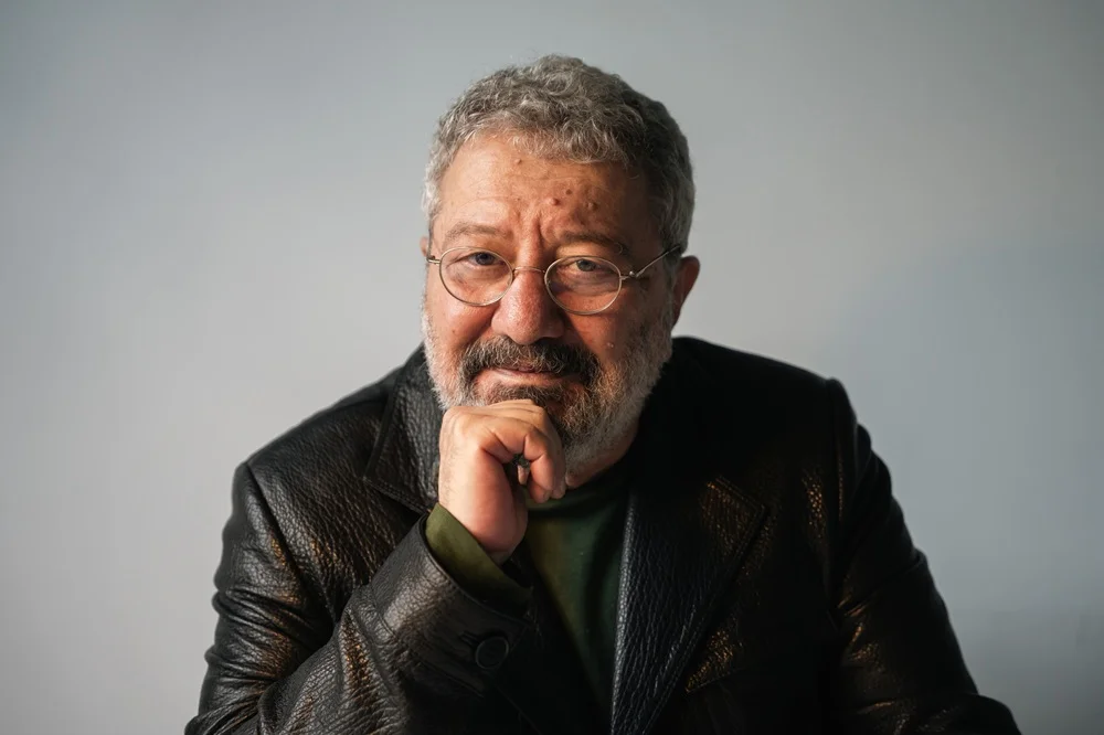 IKSV Tiyatro Festivali küratörü Mehmet Birkiye'nin, yüzünde belirgin çizgiler, beyazlamış saçları ve sakalıyla, koyu renk şık bir ceket ve açık renk bir gömlek giymiş olarak, net ve sade bir arka plan önünde düşünceli ama samimi bir ifadeyle kameraya baktığı etkileyici bir portre fotoğrafı.