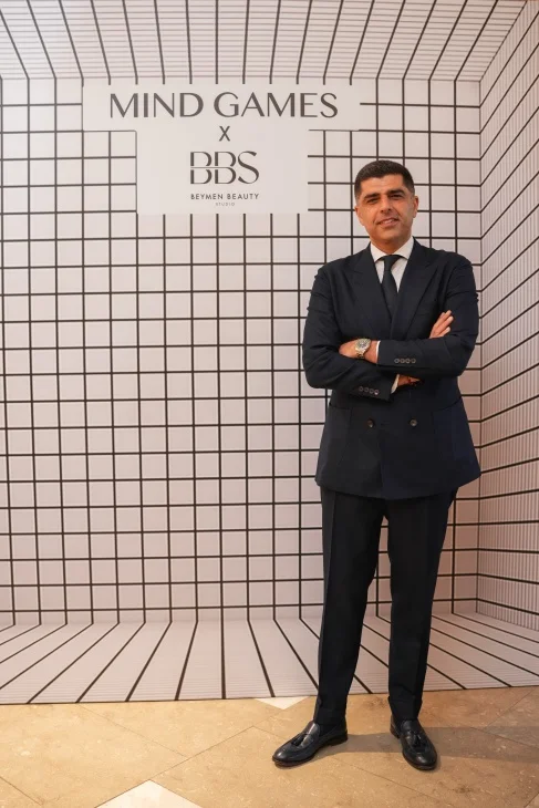 Mind Games Parfüm kurucusu Alex Shalbaf, şık bir takım elbiseyle oturduğu, arkasında modern iç mekan detaylarının bulanıkça seçildiği, röportaj anından yakalanmış, düşünceli ifadesiyle objektife bakan profesyonel bir portrede yer alıyor.