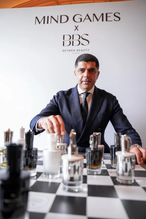 Mind Games Parfum kurucusu Alex Shalbaf, bir röportaj sırasında koyu renk takım elbisesiyle oturmuş, ellerini birleştirmiş bir şekilde hafifçe gülümserken profesyonel ve düşünceli bir duruş sergiliyor.