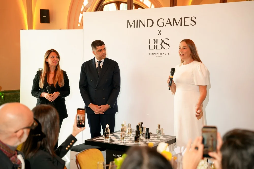 Mind Games Parfüm Kurucusu Alex Shalbaf, şık koyu renk takım elbisesiyle modern bir ofis masasında oturmuş, elleri kenetli bir şekilde kameraya sıcak bir ifadeyle bakarak gülümserken, röportajı için poz veriyor.