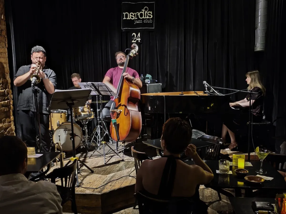 İstanbul'un en iyi caz kulüpleri arasında sayılan Nardis Jazz Club'da, karanlık sahnedeki dört kişilik bir caz grubunun trompet, kontrbas, davul ve piyano eşliğinde sergilediği büyüleyici canlı performans, izleyicilere unutulmaz bir müzik ziyafeti sunuyor.