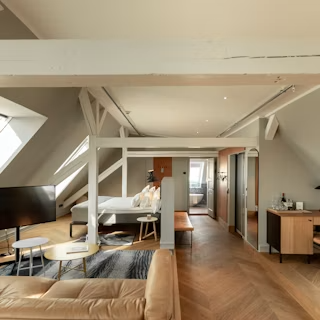 Kopenhag'ın unutulmaz otellerinden Nobis Hotel Copenhagen'da, İskandinav şıklığının ve iyi düşünülmüş tasarımın öne çıktığı geniş ve aydınlık bir süit: V desenli ahşap parke zeminleri, doğal ışığı içeri alan eğimli çatı pencereleri, beyaz tavan kirişleriyle çevrili yatak alanı, bej renkli konforlu bir kanepe, minimal sehpalar ve zarif ahşap mobilyalarla sakin bir konaklama ortamı sunuluyor.